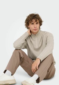 Junger Mann mit lockigem Haar, der einen beigefarbenen Kabelstrickpullover, braune Hose, weiße Socken und weiße Sneaker trägt. Er sitzt auf dem Boden und hat eine Hand um den Hals.