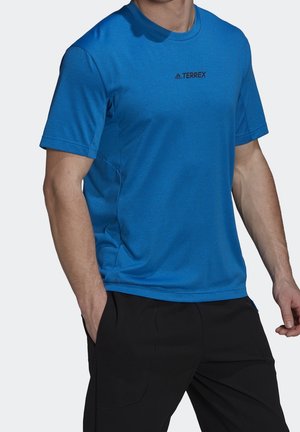 Homme portant un t-shirt bleu Adidas Terrex à manches courtes et un pantalon noir, une main dans la poche, avec un fond neutre.
