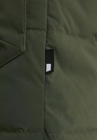 Tessuto di giacca imbottita verde oliva con cuciture visibili, con etichetta nera e bianca sul lato. Texture liscia, design isolante.