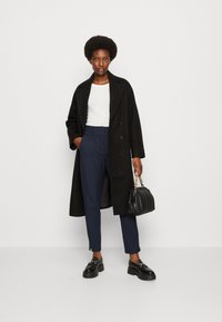 Cappotto lungo nero con ampi rever, indossato su una maglietta bianca, pantaloni blu navy e scarpe con zeppa nere. Porta una borsa a pieghe nera.
