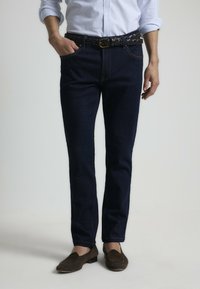 Jeans de mezclilla azul oscuro con un corte recto, que presentan costuras visibles, un cinturón de cuero trenzado y bolsillos frontales. Se llevan mocasines marrones.