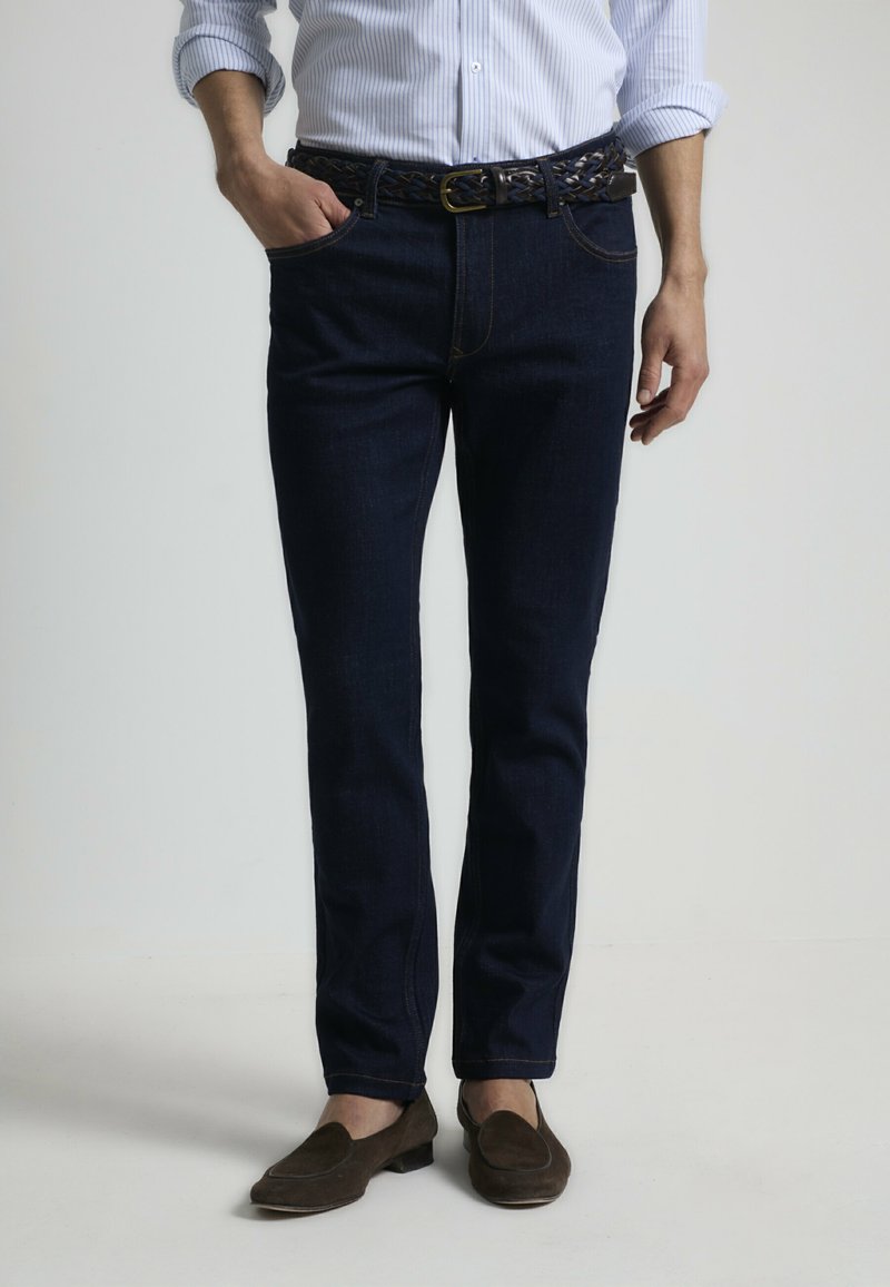 Jeans de mezclilla azul oscuro con un corte recto, que presentan costuras visibles, un cinturón de cuero trenzado y bolsillos frontales. Se llevan mocasines marrones.