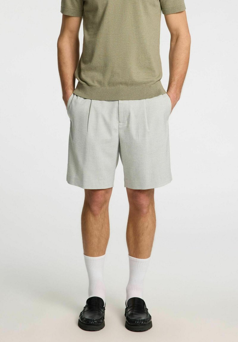Shorts plissés gris avec une texture lisse, associés à un haut en tricot à manches courtes de couleur vert olive, des chaussettes blanches et des mocassins noirs.