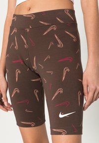 Pantalones cortos de ciclismo marrones con patrones de esbozo multicolores en rosa y naranja. Presenta un logo blanco de Nike en la pierna izquierda y un diseño ajustado.