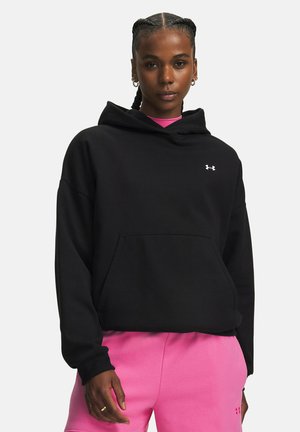 Vrouw met gevlochten haar die een zwarte hoodie en roze joggingbroek draagt met kleine Under Armour-logo's op beide kledingstukken.