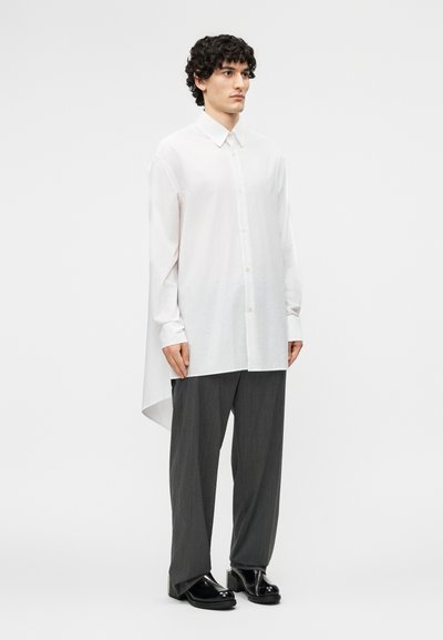 Junger Mann mit lockigem Haar trägt ein langes weißes Hemd mit Knöpfen, lockere graue Nadelstreifenhosen und schwarze Plateauschuhe, steht vor weißem Hintergrund.