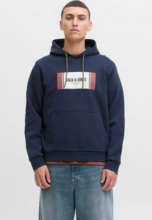 Mørkeblå hættejakke med logo-patch, snoretræk i hætten, frontlomme og ribbede manchetter. Bæret over en rustfarvet skjorte og lyse jeans.
