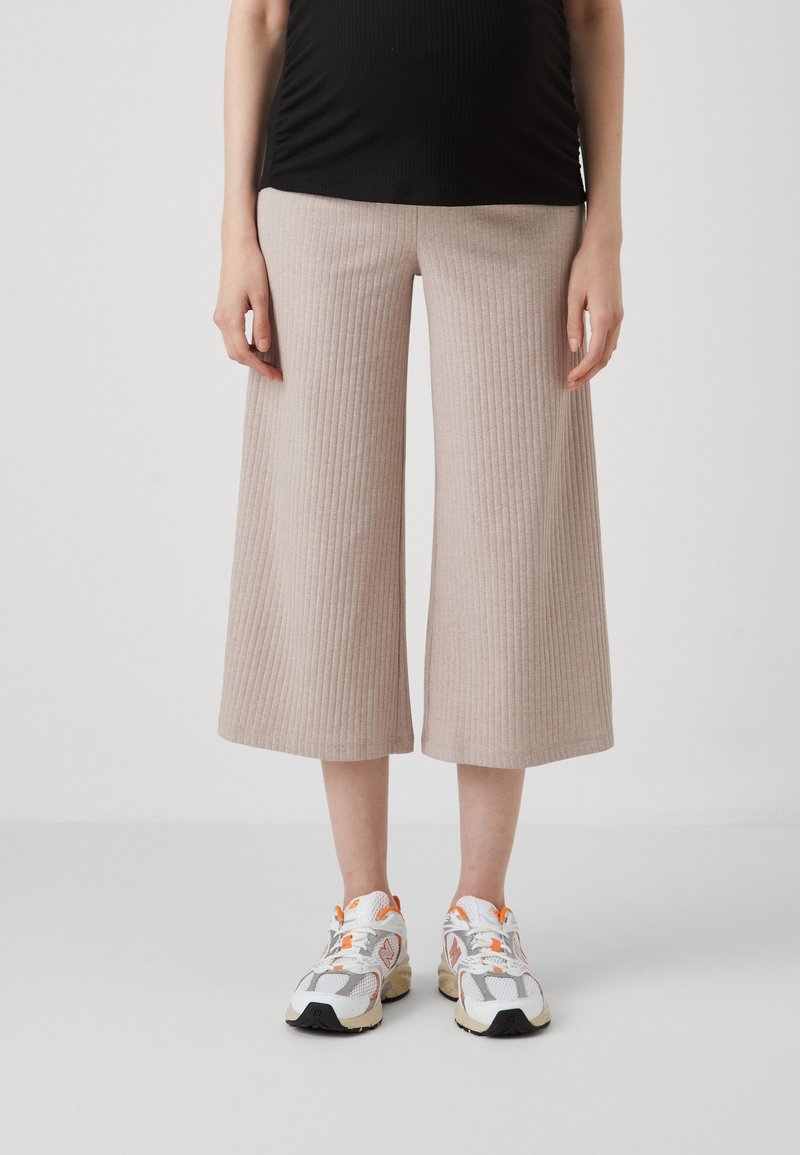 Pantalons larges côtelés beige avec une coupe décontractée, présentant une texture lisse. Associés à des baskets blanches et grises avec des accents orange.