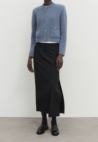 Cardigan en cachemire bleu clair court avec deux poches avant, jupe midi noire avec fente sur le côté, et mocassins noirs avec chaussettes côtelées.