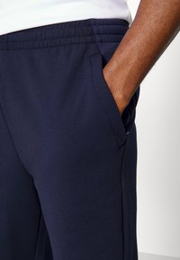 Navy urheilujuoksuhousut, joissa on sileä pinta, joustava vyötärö, sivutaskut ja verkkopaneeli yksityiskohtana yläreunoissa.