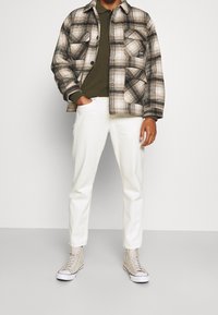 Rutiga jacka i beige och svart, olivgrön topp, vita byxor och beige högstövlar med öglor och snörning. Fyra fickor på jackan.