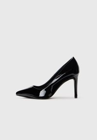 Scarpa con tacco alto in pelle verniciata nera con punta affilata, esposta su uno sfondo bianco semplice.