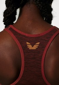 Bordeaux sporttanktop met een gestructureerd patroon, met een gouden vleugel logo op de achterkant. Racerback ontwerp met een gladde stofafwerking.