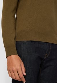Pull en tricot vert olive avec poignets et ourlet côtelés, porté avec un jean denim foncé. La texture semble lisse avec une coupe ajustée.