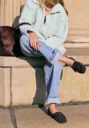 Persoană așezată pe trepte de piatră purtând blugi albastru deschis suflecați, pulover pufos verde mentă, mocasini închiși la culoare cu ciucuri și o geantă mare maro țesută.