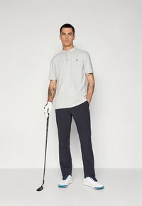 Muž v svetlosivej polokošeli, tmavých nohaviciach a bielej golfovej rukavici, drží golfovú palicu, stojí pred jednoduchým pozadím s jednou rukou v vrecku.