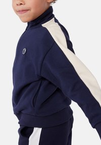 Navyblaue zip-up Jacke aus weichem Stoff, mit cremefarbenen Seitenpanelen und einem kleinen Logo auf der Brust. Hoher Kragen und gerippte Bündchen.