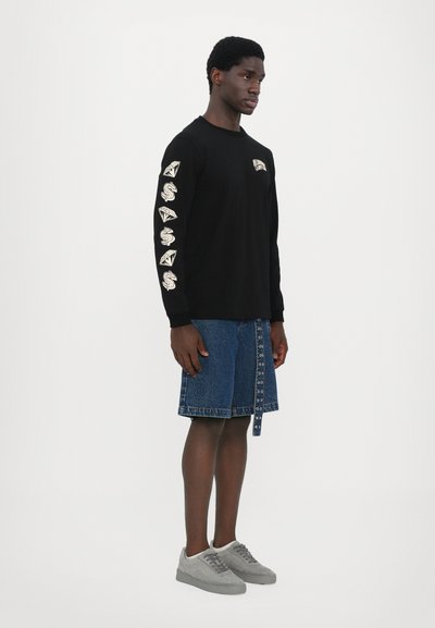 Sort langærmet t-shirt med trykte designs på ærmerne, parret med denimshorts med knapdetaljer og grå sneakers.