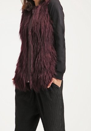 Personne portant un long gilet en fausse fourrure violet foncé sur un top noir à manches longues avec un pantalon noir texturé et ample, les mains dans les poches.