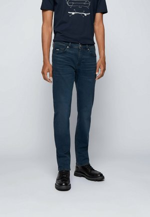 Mies, jolla on päällään tummansiniset farkut, mustat nyörikengät ja laivastonsininen t-paita, jossa on skeittilaudan kuva, seisoo yksinkertaisen vaalean taustan edessä.