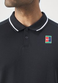 Gros plan d'une personne portant un polo noir avec des rayures blanches sur le col et un petit logo Nike multicolore sur la poitrine.