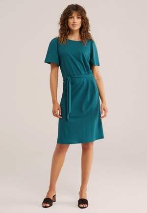 Femme debout portant une robe sarcelle mi-longue à manches courtes et ceinture à la taille, associée à des talons ouverts noirs sur un fond neutre.