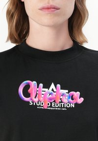 Musta lyhythihainen t-paita, jossa on monivärinen, kiiltävä muovinen "Alpha"-logo ja sen alla valkoinen "Studio Edition" -teksti.