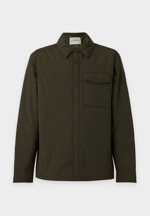 Homme debout portant une chemise boutonnée vert foncé avec une poche poitrine, un jean large bleu clair et des chaussures noires épaisses sur fond blanc.