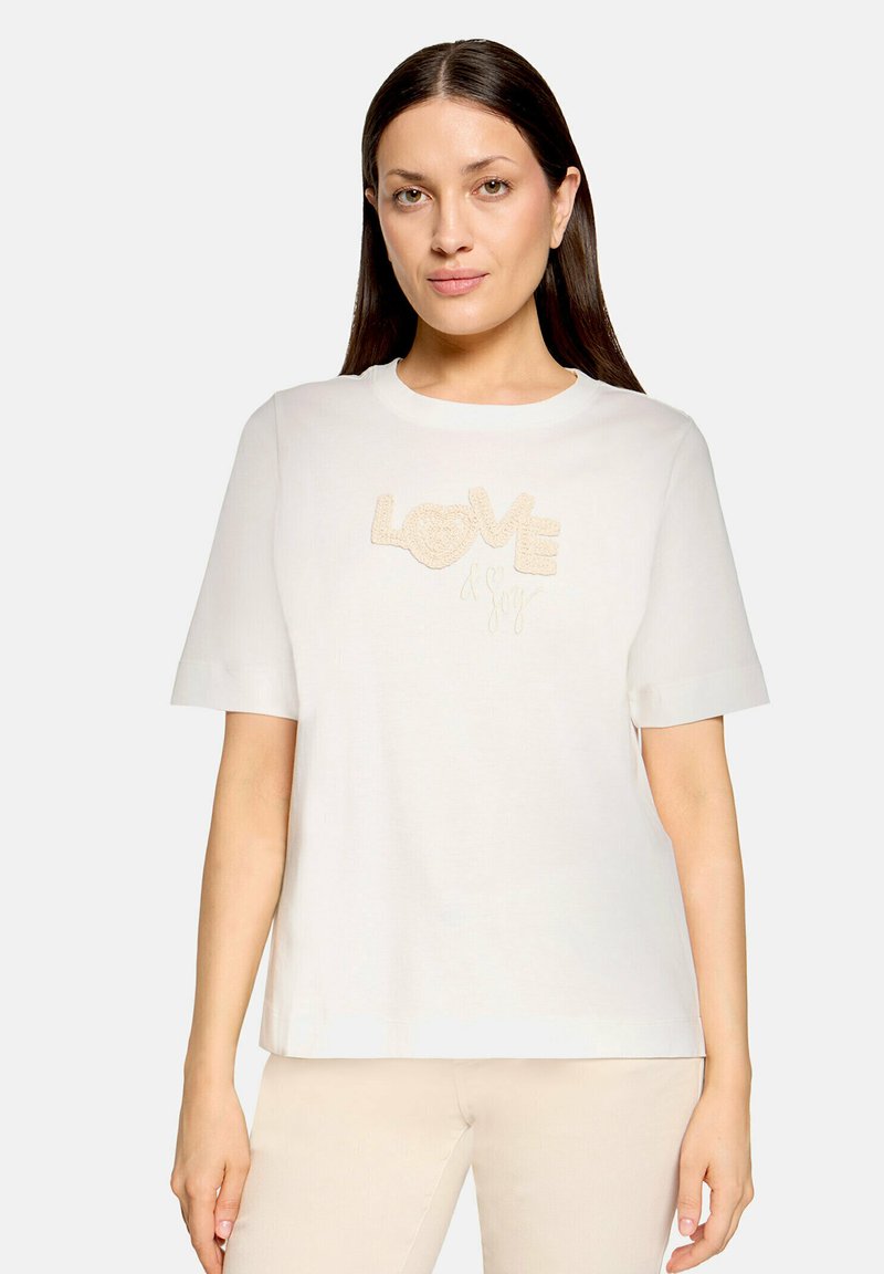 Femme aux longs cheveux bruns portant un t-shirt blanc à manches courtes avec l'inscription "LOVE & Joy" brodée en beige sur le devant.