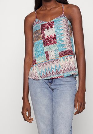 Vrouw draagt een kleurrijk mouwloos topje met zigzagpatroon en dunne bandjes, samen met lichtblauwe spijkerbroek, staand tegen een effen achtergrond.