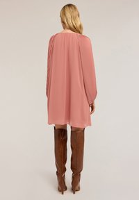 Una donna con lunghi capelli biondi indossa un abito rosa a maniche plissettate e stivali in pelle marrone al ginocchio, in piedi contro uno sfondo neutro.