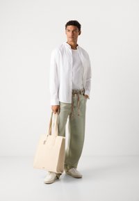 Beige tote bag with a structured shape, canvas material, dual top handles, and a front pocket. Model wears white shirt and light green pants.