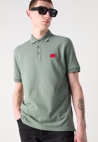 Grønn piké med klassisk krage, knappestolpe, ribbede ermer og et rødt logo-patch. Modellen har på seg svarte solbriller.