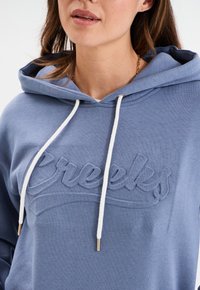Felpa blu con cappuccio caratterizzata da un logo in rilievo "Creeks", cordini bianchi e dettagli metallici dorati. Tessuto morbido con una vestibilità rilassata.