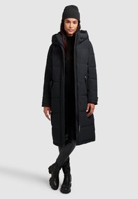 Abrigo de plumas negro con una silueta larga, diseño acolchado, capucha ajustable y bolsillos laterales, acompañado de un gorro de punto negro.