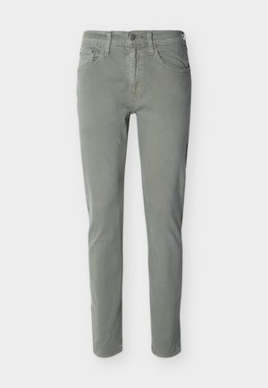 Schmale, lässige Herrenhose in gedämpftem Grün mit Knopf vorne, Reißverschluss und Fünf-Taschen-Design vor einfarbigem Hintergrund.