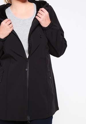 Person iført en sort lynlåssweatshirt med hætte over en grå skjorte, holder jakken åben i brysthøjde mod en hvid baggrund.