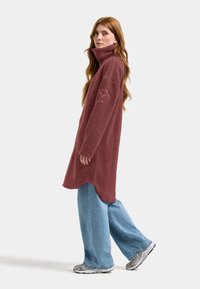 Bordeaux fleece pullover met hoge kraag, voorzien van lange mouwen en een ontspannen pasvorm, gecombineerd met lichtblauwe wijde jeans en sportieve schoenen.