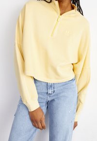 Keltainen lyhyt collegepaita, jossa on neljännesvetoketjukaulus, pitkät hihat ja kohopainettu logo rinnassa, yhdistetty vaaleansinisiin farkkuihin.