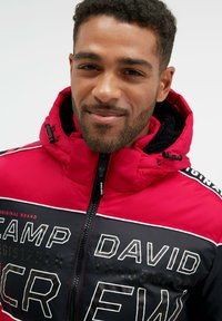 Rot schwarze Zip-Jacke mit Kapuze, ausgestattet mit auffälliger weißer Schrift und horizontalen Streifen. Strukturiertes Material mit sichtbaren Reißverschlüssen und Akzenten.