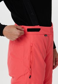 Koraal ski-pantalons hebben verstelbare zwarte banden, een ritssluitingszak aan de zijkant en een gladde textuur met twee knoopsluitingen op de taille.