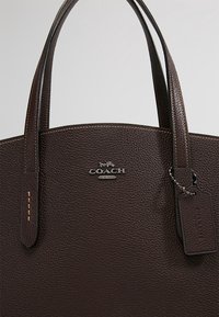 Sac à main en cuir marron avec une finition texturée, deux poignées et un logo argenté brillant. Comprend une étiquette de bagage et des surpiqûres contrastées.