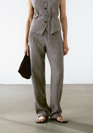 Woman wearing gray buttoned sleeveless top and matching wide-leg trousers, holding a black clutch and wearing black sandals with toe rings.