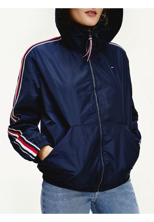 Femme portant une veste à capuche bleu marine avec des bandes rouges, blanches et bleues sur les manches, mains dans les poches, associée à un jean bleu clair.