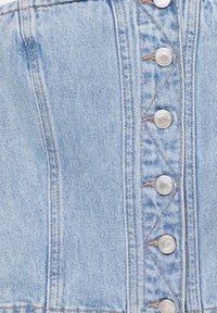 Haut en denim bleu clair avec un devant à boutons, présentant des boutons en métal argenté, des coutures contrastantes orange et un design ajusté. La texture est lisse.