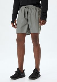 Ljusgråa träningsshorts med elastiskt midjeband och svart dragsko, med en slät yta och knälång skärning. Bärs med svarta skor.