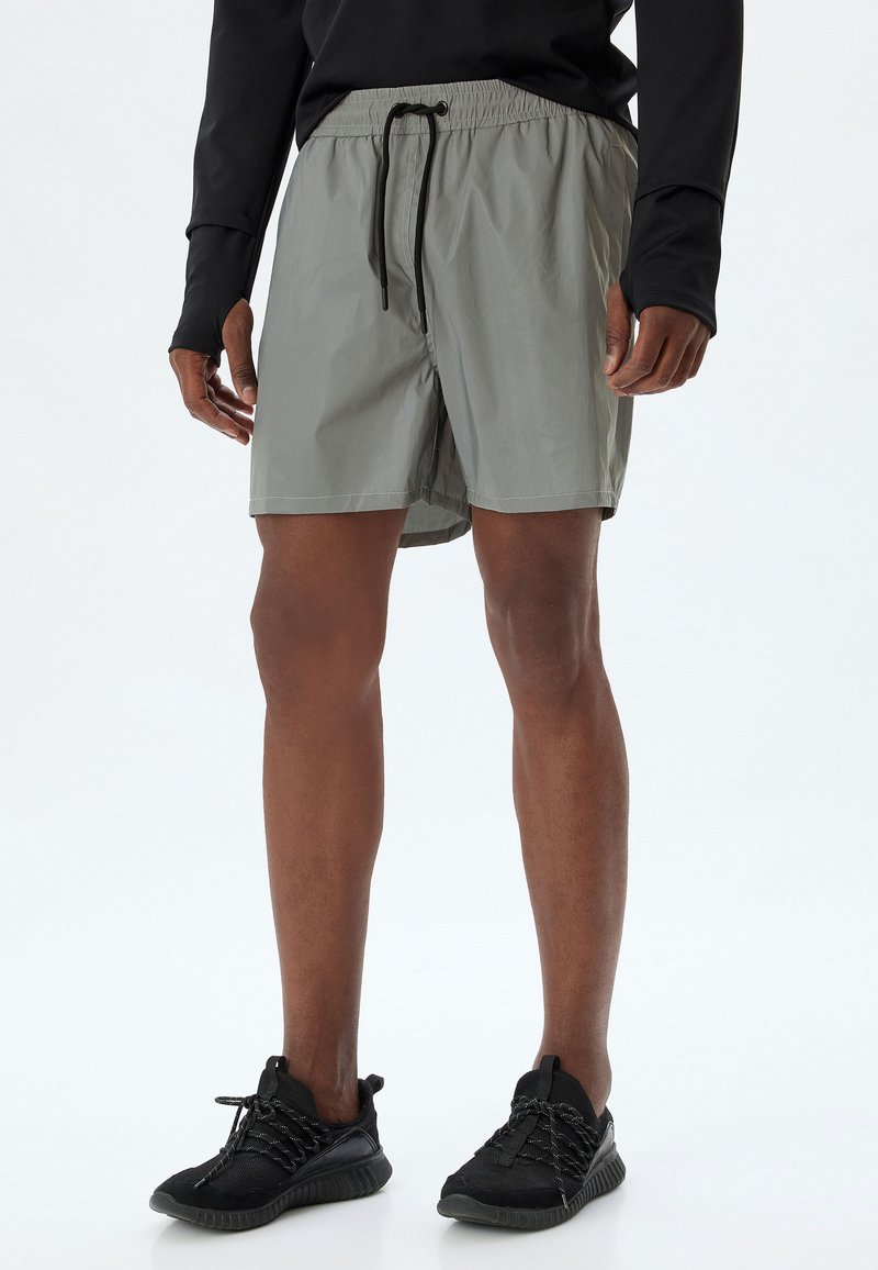 Ljusgråa träningsshorts med elastiskt midjeband och svart dragsko, med en slät yta och knälång skärning. Bärs med svarta skor.