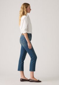 Balta mygtukų marškiniai, mėlyni vidutinio liemens, trumpi džinsai, tekstūruoti džinsai ir rudais plokščiais bateliais. Šoninis asmens stovėjimo vaizdas.