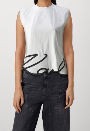 Frau trägt ein ärmelloses weißes Oberteil mit schwarzem kursivem Text nahe dem Saum und dunkle High-Waist-Jeans mit großen Fronttaschen.