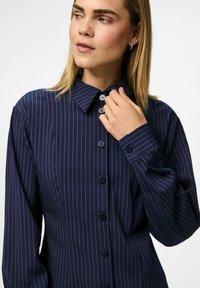 Woman with straight blonde hair wearing a navy blue buttoned shirt with thin red vertical stripes, adjusting the collar with her right hand.